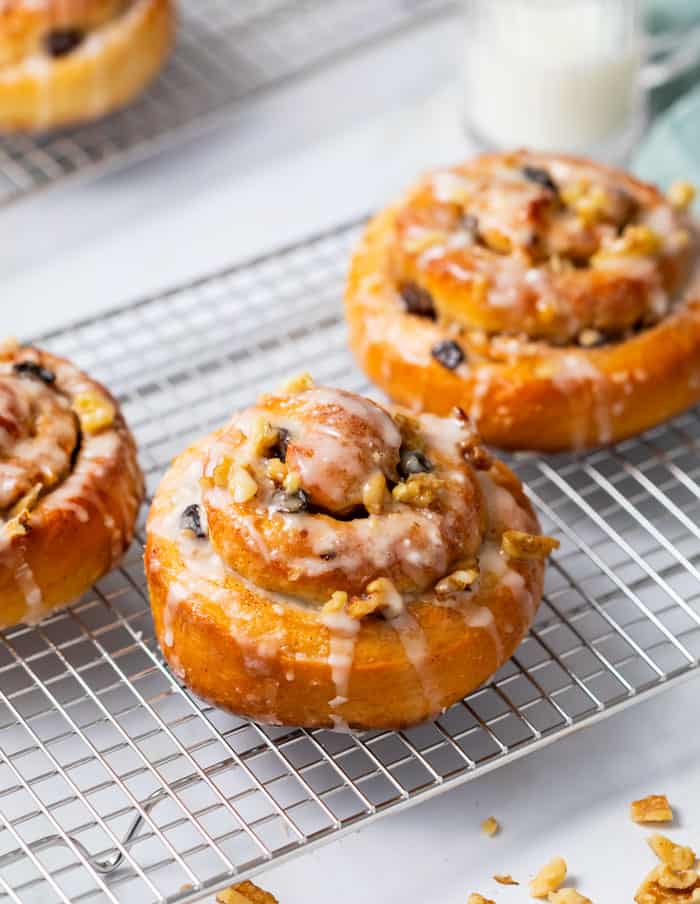 This easy Cinnamon Rolls recipe is from my grandmother, and is the best part about Thanksgiving and Christmas! A perfect holiday breakfast idea for the family. #brunch #sweetrolls #rolls #holidaybrunch #holidaybreakfastideas #makeaheadbreakfastideas Homemade cinnamon rolls on a wire rack topping with raisins and white icing.