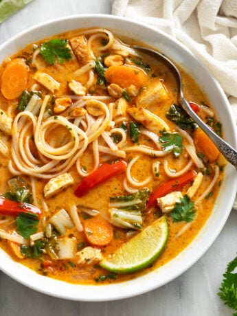 Thai Chicken Soup in a white bowl with noodles, vegetables, chicken, and a spoon and a lime wedge on the side.